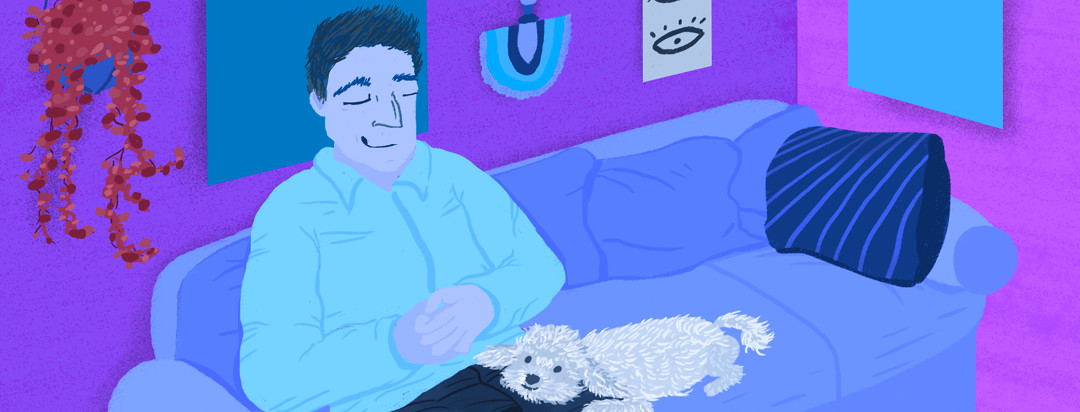 A man sitting on a living room couch with his hands folded and a smile on his face. His smiling dog rests his head on his owner's leg.