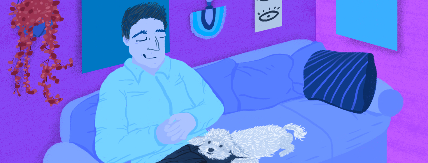 A man sitting on a living room couch with his hands folded and a smile on his face. His smiling dog rests his head on his owner's leg.