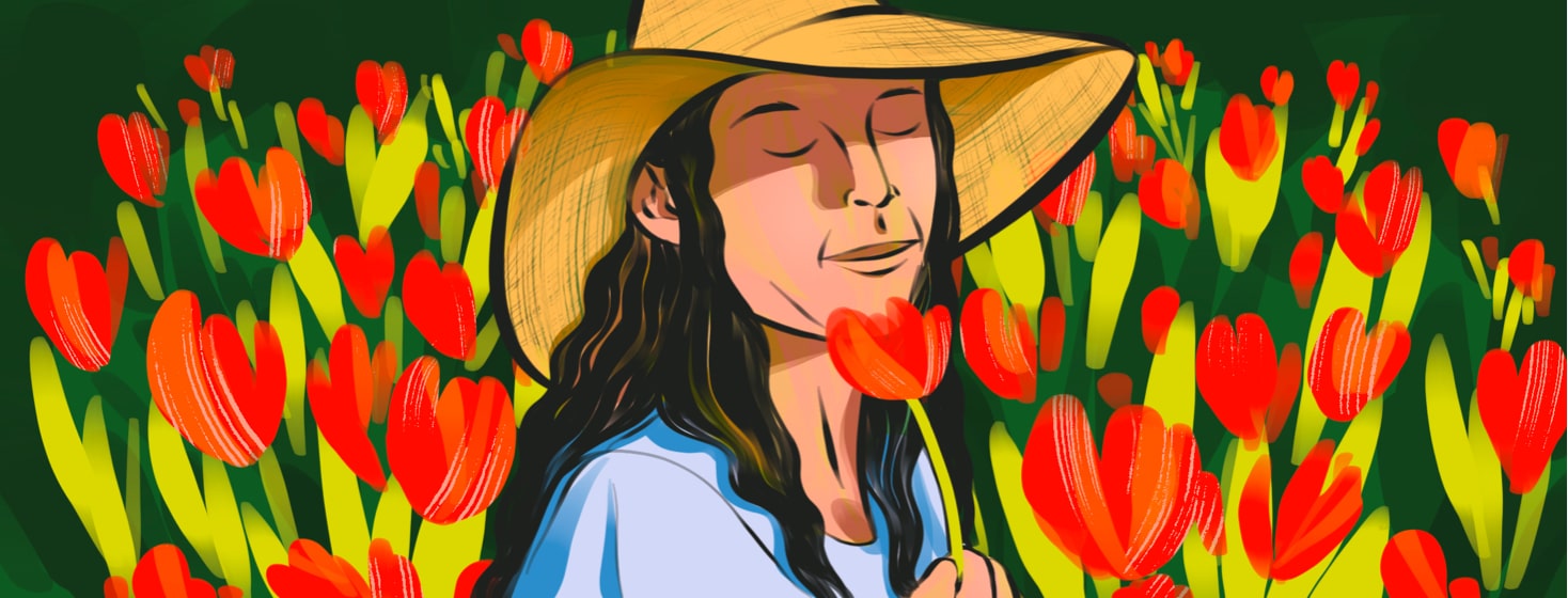 alt=A woman in a sunhat closes her eyes and smells a flower she is holding, while standing in a field of red flowers.