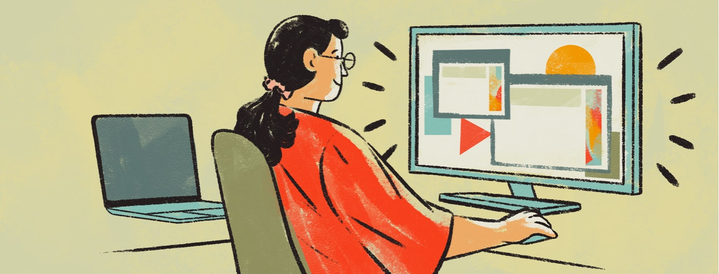 alt=a woman sitting at a desk smiles at a large active computer monitor. A small dark laptop is open to her left.